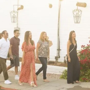 A group of eight people walk along a sidewalk next to a white stucco building with hanging lanterns and red flowering bushes.