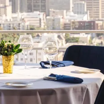 A round table set with wine glasses, plates, and blue napkins overlooks a cityscape through large windows; dark blue chairs surround the table.