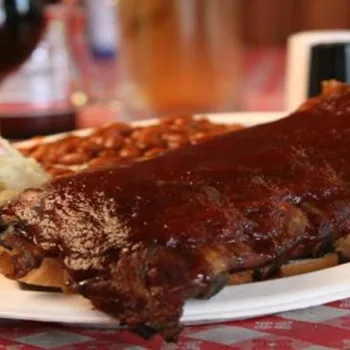 A plate with barbecue ribs on bread, baked beans, and potato salad on a red and white checkered tablecloth.