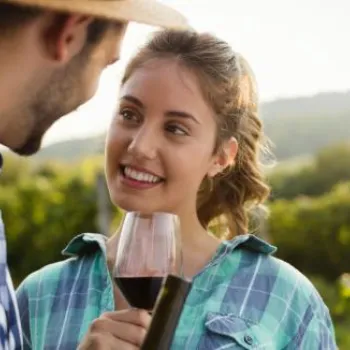 A woman holding a glass of red wine smiles at a man wearing a hat in a vineyard.