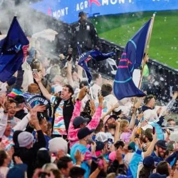 Crowd of soccer fans cheering and waving flags in a stadium, with confetti in the air and a security guard standing nearby.