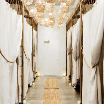 A hallway with white fabric curtains on both sides, wicker light fixtures on the ceiling, and a woven rug runner on the concrete floor, leading to a closed white door.