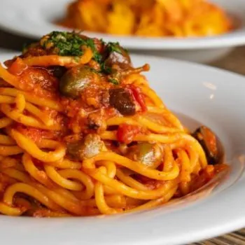 A plate of spaghetti with tomato sauce, garnished with herbs, sits on a white dish with other plates of food visible in the background.