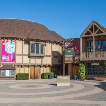 A round, brown, Tudor-style theater building sits beside a modern, angular building under a clear blue sky; banners and outdoor seating are visible.