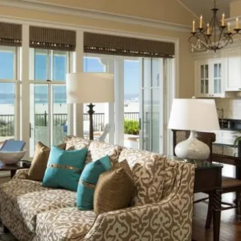 Bright living room with patterned sofa, kitchen area, and large windows offering a view of a beach and ocean.
