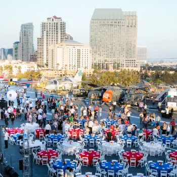 People gather around tables set with red, white, and blue tablecloths on the deck of a ship with helicopters and city buildings in the background.