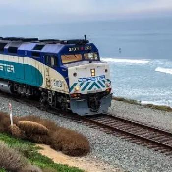 A Coaster commuter train travels along tracks beside the ocean, with waves in the background and greenery on the left side.