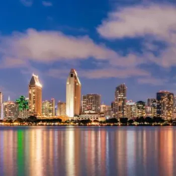 San Diego city skyline at dusk, with illuminated buildings reflected in calm water under a partly cloudy sky.