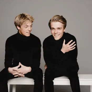 Two young men in black turtlenecks sit on a white bench against a plain gray background. One is smiling at the camera while the other looks at him and gestures with his hands.