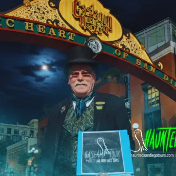A man in Victorian attire stands under the Gaslamp Quarter arch in San Diego at night, holding a sign for haunted San Diego ghost tours.