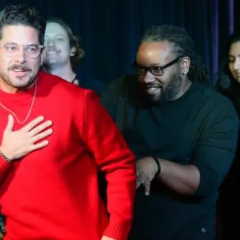 A man in a red sweater stands in front, surrounded by a group of people smiling and interacting on a dark stage.