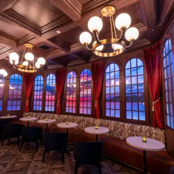An elegant lounge with round tables, cushioned benches, dark chairs, large arched windows, red curtains, and ornate chandeliers under a wood-paneled ceiling.