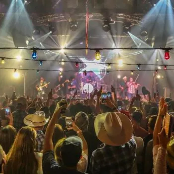A large crowd attends a lively concert in an indoor venue, with people raising their hands and a band performing on a brightly lit stage.