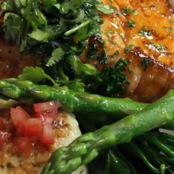 Grilled salmon fillet topped with herbs, served with asparagus, broccolini, and a potato cake garnished with diced tomatoes on a white plate.