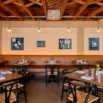 A modern restaurant dining area with wooden tables, black chairs, a long brown bench, small vases with flowers, and framed artwork on the walls under exposed wooden ceiling beams.
