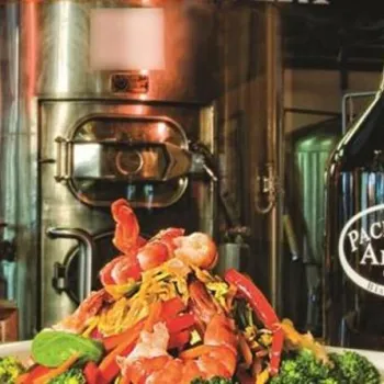 A plate of shrimp and vegetables sits next to a glass of beer and a Pacific Beach Alehouse growler, with brewing equipment in the background.