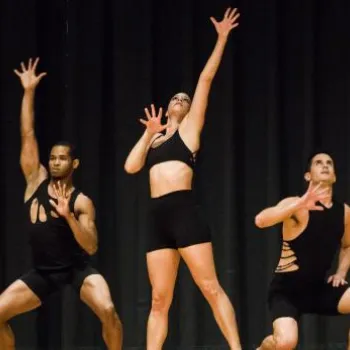 Five dancers in black outfits perform a choreographed routine on stage, each striking dynamic poses with arms raised against a black curtain backdrop.