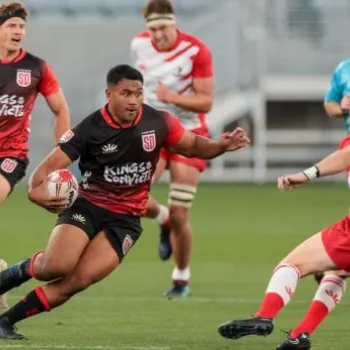 A rugby player in a black and red uniform runs with the ball while players in red attempt to block him during a match on a stadium field.