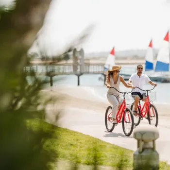 Two people ride red bicycles along a waterfront path, with sailboats and a pier visible in the background on a sunny day.
