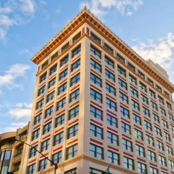 A multi-story historic building with decorative trim stands on a city corner under a partly cloudy sky, surrounded by other modern buildings.