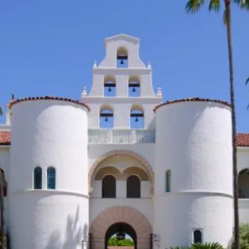 A white stucco building with red tile roof, twin towers, arched entrance, and a bell tower, surrounded by palm trees under a clear blue sky.