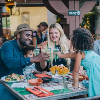 A family of four sits at an outdoor restaurant table, clinking glasses and smiling, with food and drinks on the colorful tiled table.