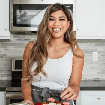 Woman in a white tank top and apron smiles while preparing food in a modern kitchen with white cabinets and stainless steel appliances.