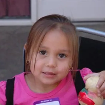 A young girl in a pink shirt sits on a chair and holds up a painted rubber duck, looking at the camera.