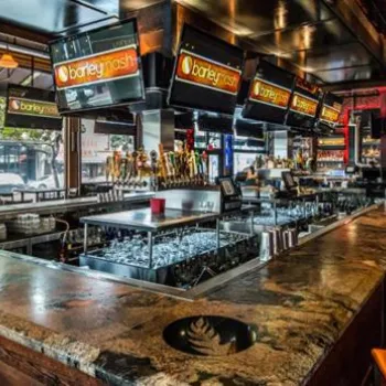 A spacious bar with high chairs, multiple beer taps, overhead TVs, and large windows providing street views in a modern, industrial-style setting.