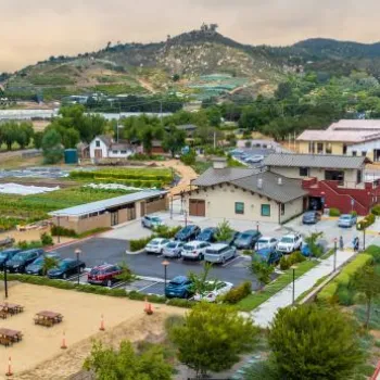 Aerial view of a community farm and garden area with parked cars, a fitness area, several buildings, and hills in the background under a cloudy sky.