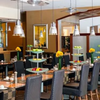 Modern restaurant dining area with black woven chairs, wooden tables, tiered trays with fruit and flowers, pendant lights, and a view of the open kitchen and chalkboard menu on the left.