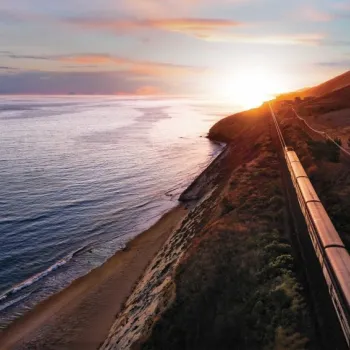 A train travels along a coastal hillside at sunset, with the ocean on one side and a sandy beach below.