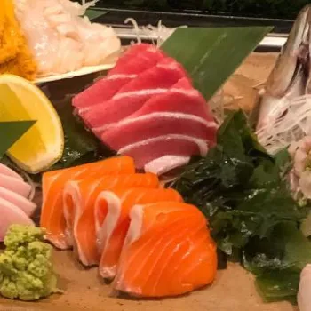 A platter of assorted sashimi, including slices of tuna, salmon, white fish, and shellfish, with wasabi, lemon, and garnishes, served on a rectangular plate.