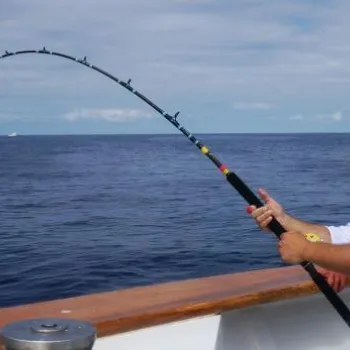 Person wearing a straw hat and sunglasses fishing on a boat, holding a bent fishing rod over the ocean on a partly cloudy day.