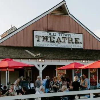 A crowd gathers under red umbrellas outside the Old Town Theatre, a rustic wooden building with a large sign and white fencing.