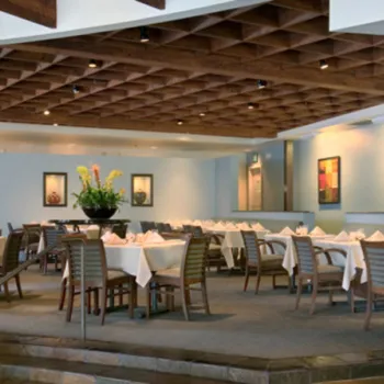 Modern restaurant interior with wooden tables and chairs, white tablecloths, framed art on pastel walls, and a decorative wooden ceiling. Green plants are placed throughout the space.