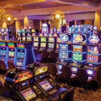 Wide view of a brightly lit casino floor filled with rows of colorful slot machines and empty chairs, under modern hanging lights.