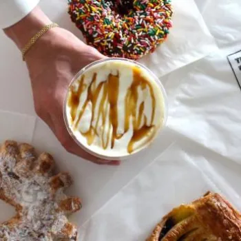 A hand holds a cup with caramel drizzle over whipped cream, surrounded by a sprinkled donut and pastries on napkins labeled "THE MRKT.