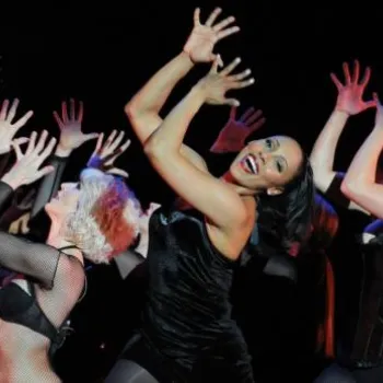 Several performers in black costumes dance on stage, striking dramatic poses with raised hands, under theatrical lighting against a dark background.