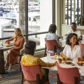 People dining at tables inside a restaurant with large windows overlooking a marina with docked boats.