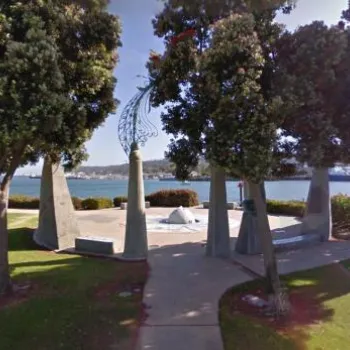 A waterfront park features a circular monument surrounded by trees, with benches facing the water and buildings visible across the bay.