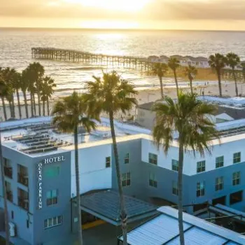 Aerial view of a beachfront hotel with palm trees, a sandy beach, a pier extending into the ocean, and the sun setting over the water.