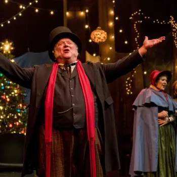 A man in Victorian costume with a red scarf sings with arms outstretched on stage; two women in period dress sing behind him, with a Christmas tree and festive lights in the background.