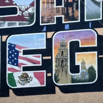 Woman in hat and sunglasses poses in front of a colorful "San Diego" mural; a bicycle is parked nearby.