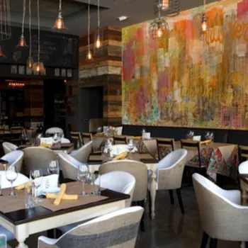 A modern restaurant dining area with neatly set tables, cushioned chairs, hanging pendant lights, and a colorful mural on the wall.