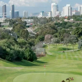 A golf course green is in the foreground with city buildings and skyscrapers in the background under a partly cloudy sky.