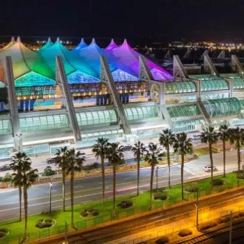 San Diego Convention Center at night, illuminated with multicolored lights on its rooftop, surrounded by palm trees and city buildings.