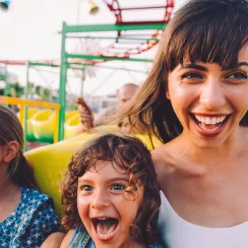 Three people, an adult and two children, are smiling and enjoying a ride on a roller coaster at an amusement park during the day.