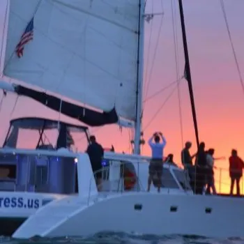 A white catamaran named Adventuress.us sails on the ocean at sunset with several people on board.