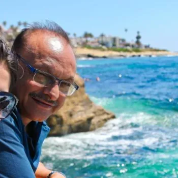 A woman and a man stand near a rocky shoreline, smiling at the camera. The blue ocean and coastal houses are visible in the background under a clear sky.